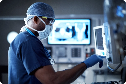 Surgeon adjusts a monitor in an operating room, wearing scrubs, surgical cap, mask, and gloves; medical equipment and monitors behind.